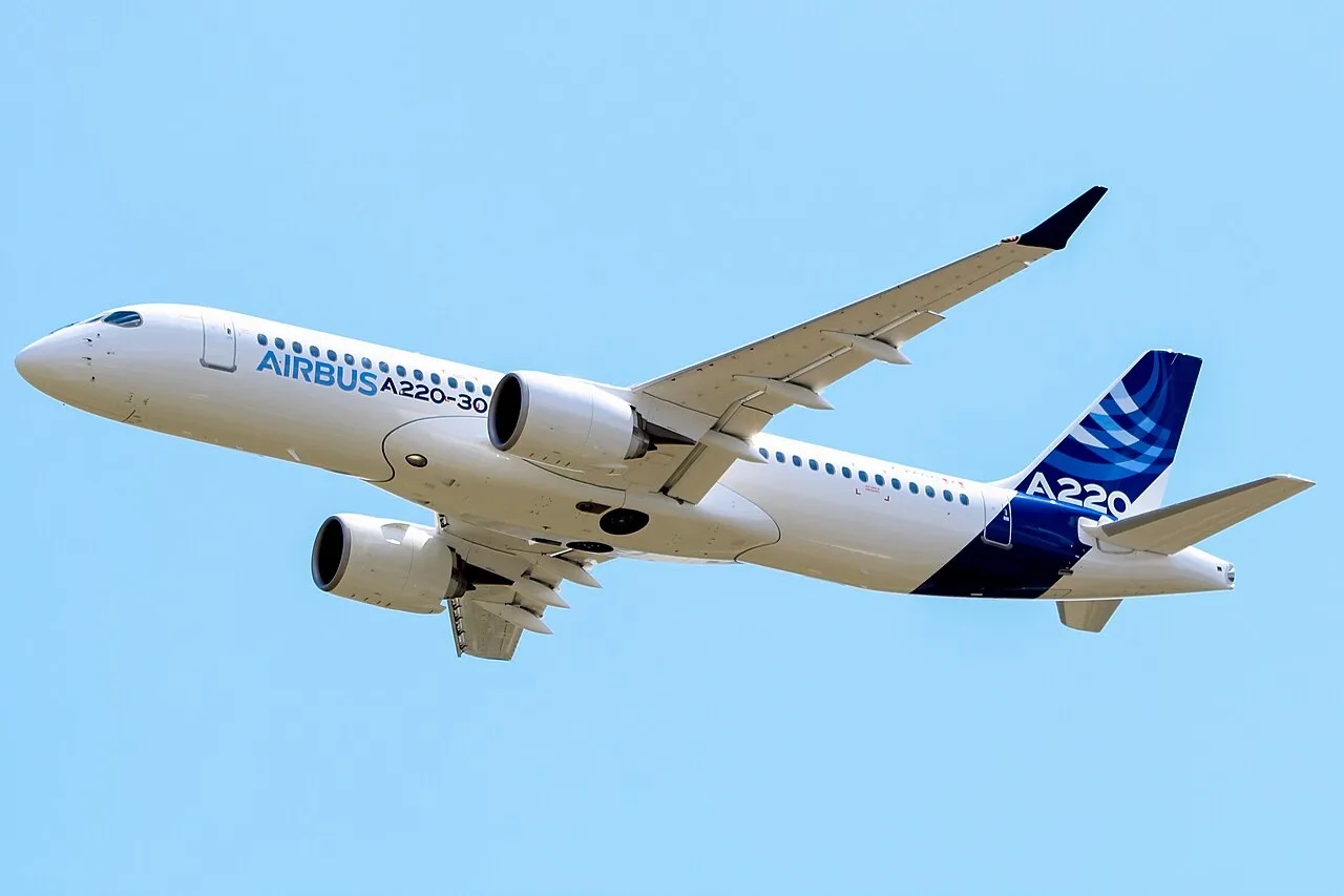 An Airbus A220-300 test aircraft in Airbus livery flying low over Toulouse-Blagnac airport.