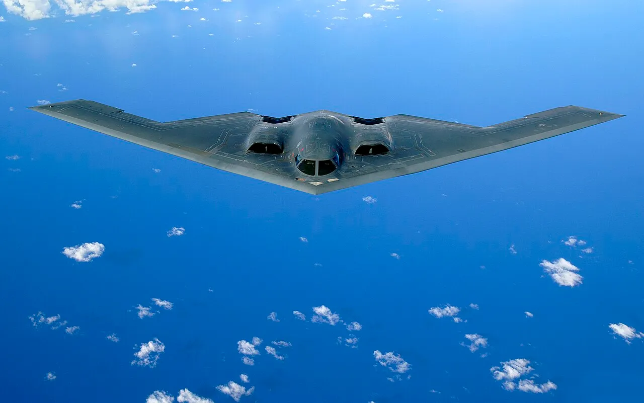 A Northrop B-2 Spirit stealth bomber, a flying wing with no tail, banking against a blue sky after a refuelling mission over the Pacific Ocean.