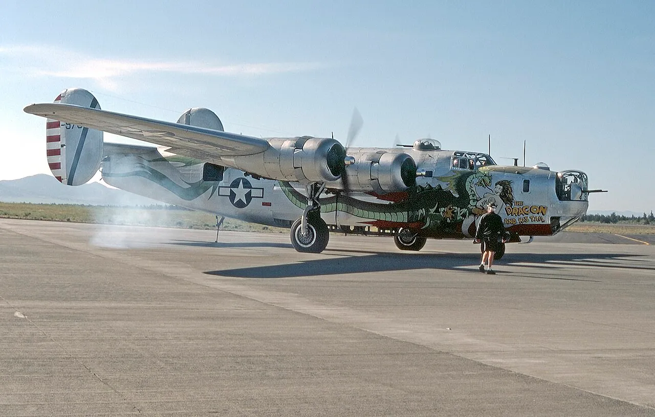 A fully restored, silver Consolidated B-24J Liberator bomber on a runway apron, nose art reading "The Dragon And His Tail" visible below the cockpit.