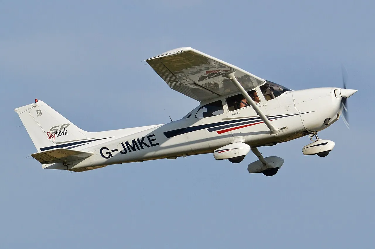 A white and blue Cessna 172S Skyhawk light aircraft seen departing from an airfield, its high wing and fixed landing gear visible.