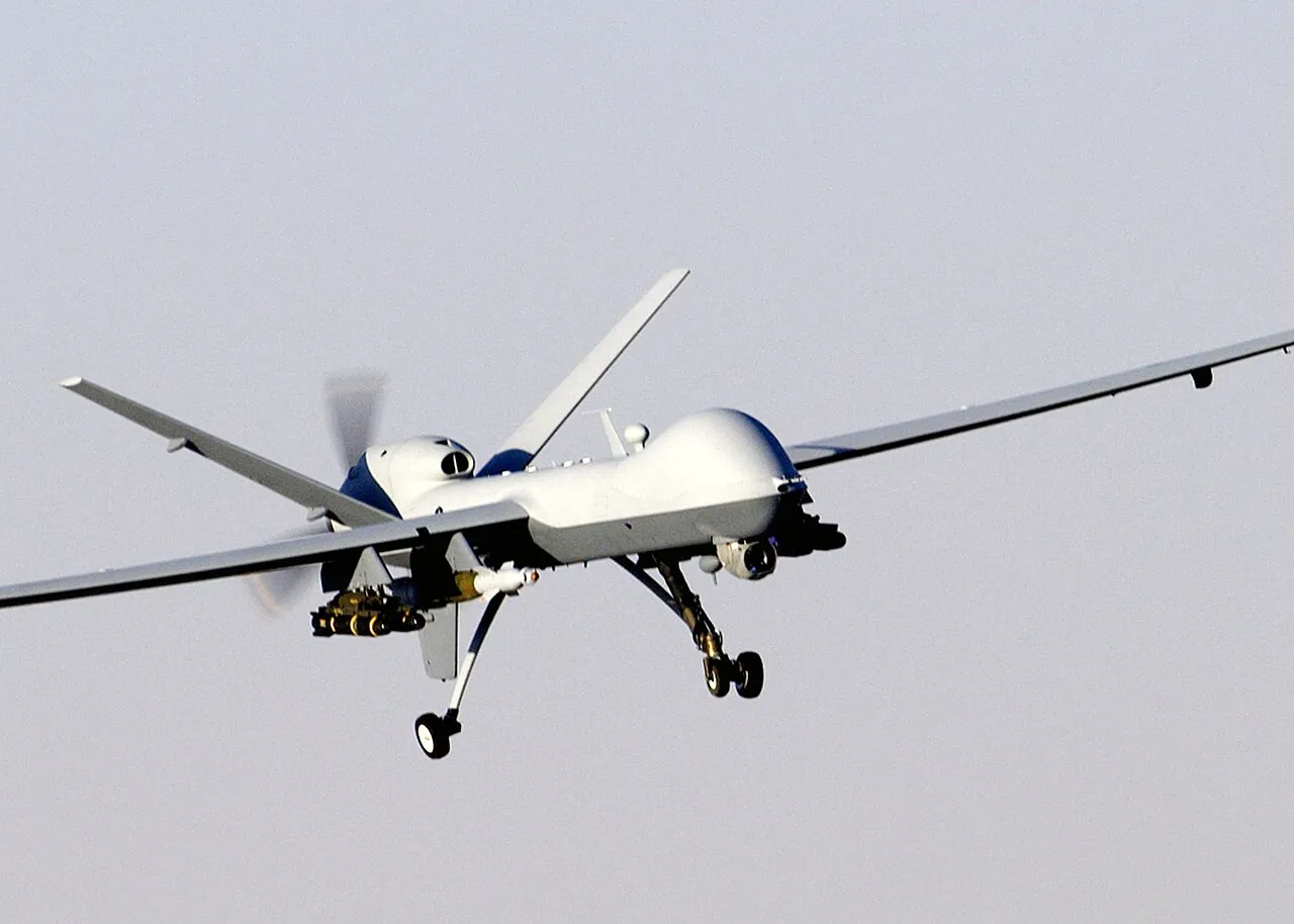 A grey MQ-9 Reaper unmanned aircraft in flight over a cloudy sky, its slender fuselage and inverted V-tail visible.